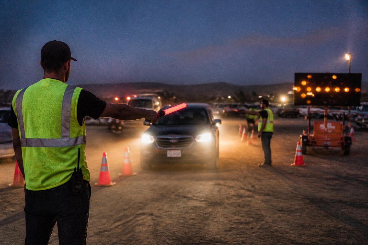 Traffic control staff in high-visibility vests directing arriving vehicles at an event parking entrance at dusk